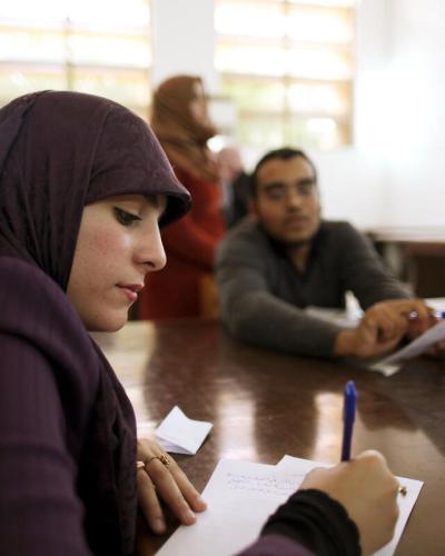 A young Libyan woman wearing a purple headscarf sits at a wooden table taking notes.